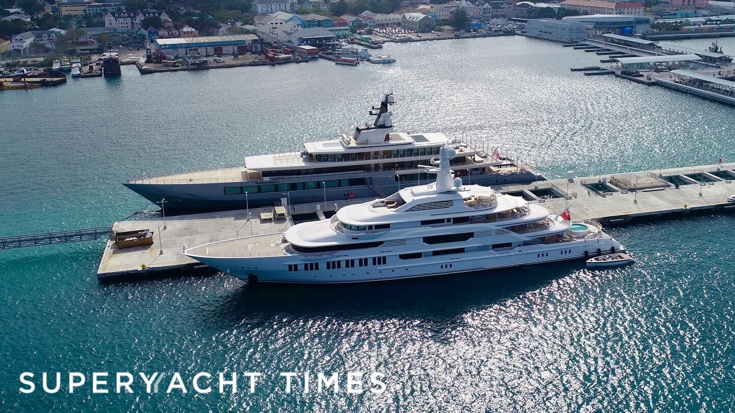 Aerial view of two luxury yachts docked at a marina, with a scenic waterfront and vibrant buildings in the background.
